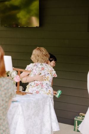 Two women in floral dresses sharing a warm hug at an outdoor wedding celebration.