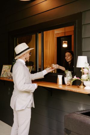 Man in white suit and hat receives a cocktail from a bartender at an elegant outdoor bar window.