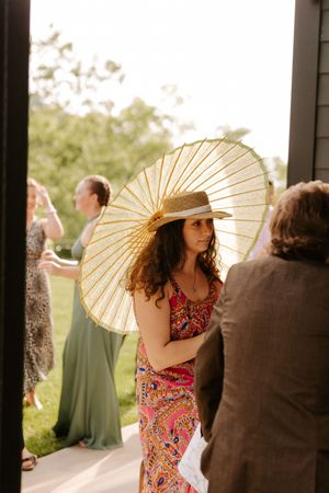 Woman in floral dress and straw hat holding a gold parasol at an outdoor summer wedding event.
