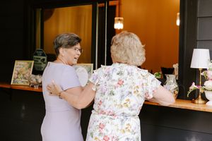 Two elderly women in dresses chat warmly near a wooden counter indoors at a social gathering.