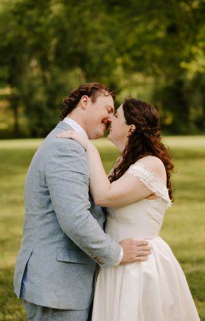 Smiling bride and groom embrace closely in lush green outdoor setting, bride in white lace off-shoulder gown and groom in blue suit.
