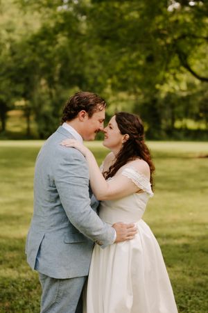 Bride and groom share an intimate nose-to-nose moment embracing in a sunlit green outdoor setting.