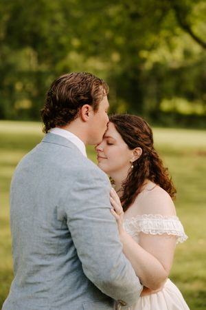 Groom tenderly kissing bride on forehead as she smiles with eyes closed in green garden.