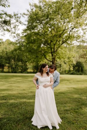 Groom embraces bride from behind as she smiles, standing on a lush green lawn beneath a large tree.