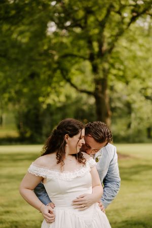 Groom tenderly embraces bride in white off-shoulder gown under large tree in green garden.