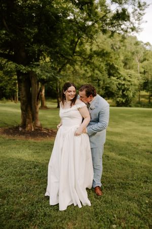 Groom in light blue suit whispering to smiling bride in white dress on a lush green lawn.