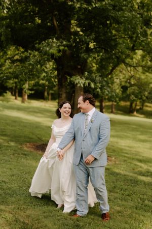 Smiling bride in white gown and groom in light blue suit walk hand in hand through a lush green park.