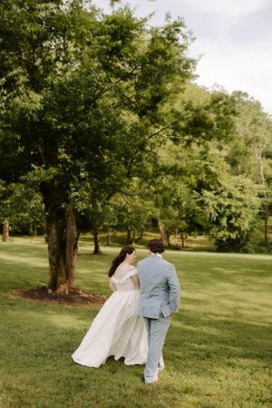 A bride in a white gown and groom in a light blue suit walk hand in hand toward trees on a sunny day.