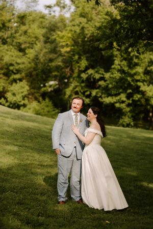 Bride in off-shoulder white gown laughs with groom in light blue suit on a green hillside wedding venue.