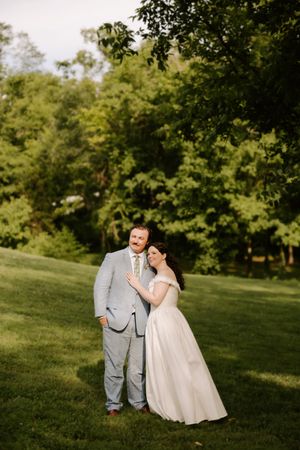 Bride in off-shoulder gown leans on groom in light gray suit posing in golden hour garden light.