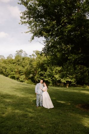 A bride and groom pose together on a lush green hillside surrounded by tall trees on their wedding day.
