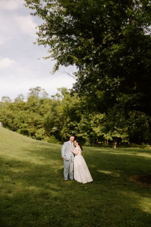 Bride and groom pose together on lush green hillside surrounded by trees at outdoor wedding.