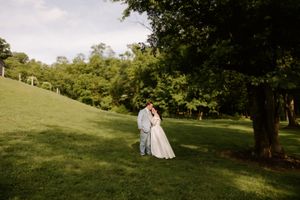 A bride and groom share a romantic kiss on a lush green hillside surrounded by tall trees on their wedding day.