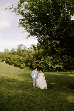 Bride and groom walking hand-in-hand across lush green lawn surrounded by tall trees on wedding day.