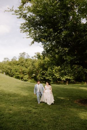 Bride and groom walk hand in hand across lush green hillside beneath large trees at wedding.