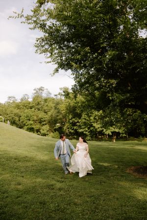 Bride and groom hold hands walking across green hillside lawn beneath large shade tree.