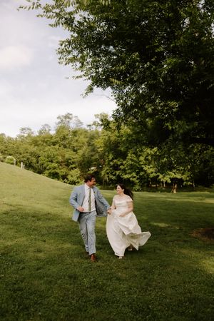 Smiling bride and groom hold hands while walking on green grass surrounded by tall trees on wedding day.