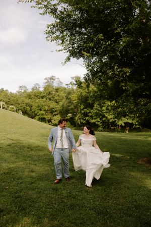 Bride and groom walk hand-in-hand across lush green hillside, bride's white gown flowing in the breeze.