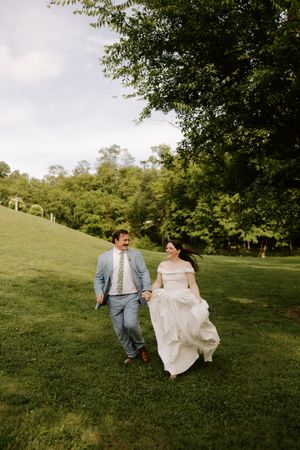 Happy bride and groom holding hands running across lush green lawn on their wedding day.