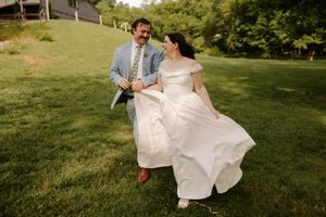 A joyful couple runs across a lush green lawn, bride in white gown and groom in blue suit, laughing together.