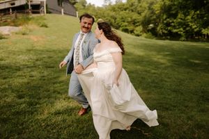 A joyful bride in a white off-shoulder gown and groom in a light blue suit run together across a lush green lawn.