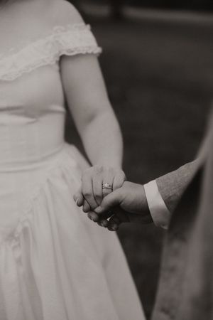 Black and white close-up of bride and groom holding hands showing wedding ring detail.