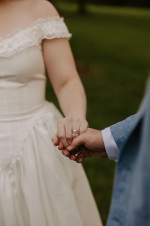 Close-up of bride and groom holding hands showing wedding ring set and off-shoulder bridal gown.