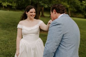 Bride in off-shoulder white gown laughs with groom in blue suit outdoors on green grass at wedding.