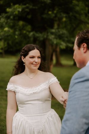 Bride in off-shoulder white gown smiles at groom during outdoor wedding ceremony in lush green park.