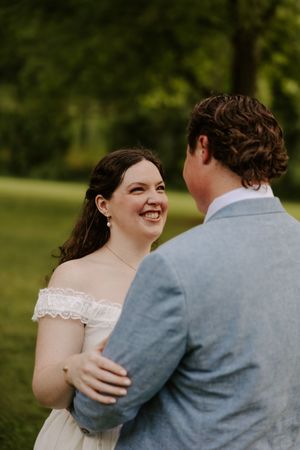 Joyful bride in lace off-shoulder wedding dress laughs with groom in blue suit outdoors.