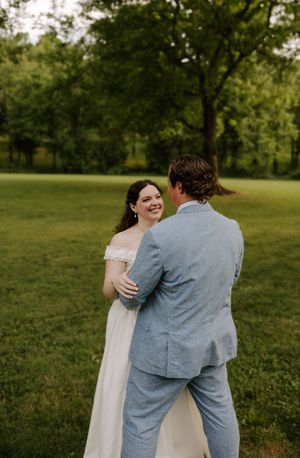 Smiling bride in white off-shoulder gown dances with groom in light blue suit on grass.