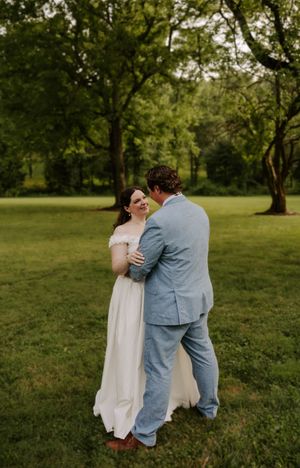 Groom in light blue suit embracing bride in off-shoulder white gown on green lawn.