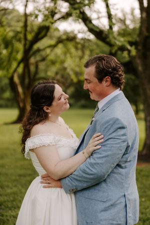 Bride in off-shoulder white gown and groom in light blue suit embrace tenderly in lush green garden.