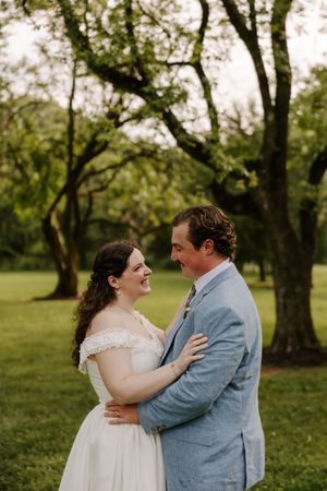 Smiling bride in white off-shoulder gown embraces groom in blue suit outdoors at wedding.