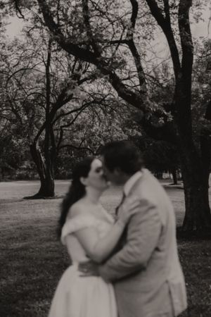 Black and white romantic photo of bride and groom sharing a kiss under trees outdoors.