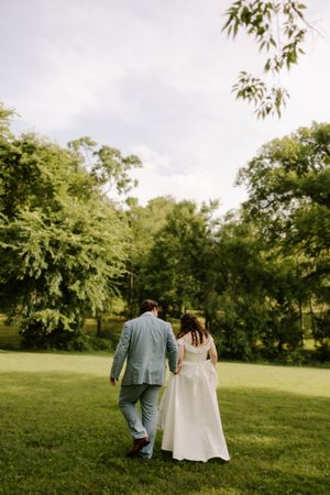 Bride and groom walk hand in hand away from camera through green trees at outdoor wedding.