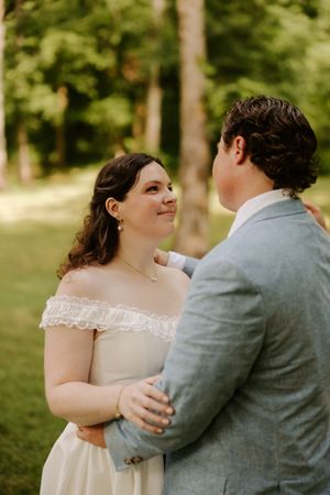 Bride in off-shoulder white gown gazes lovingly at groom in light blue suit outdoors among green trees.