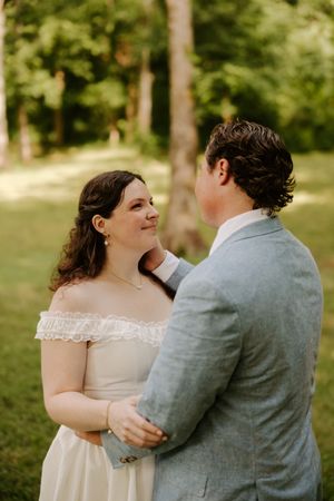 Bride smiles warmly at groom while embracing in a sunlit wooded garden setting on their wedding day.