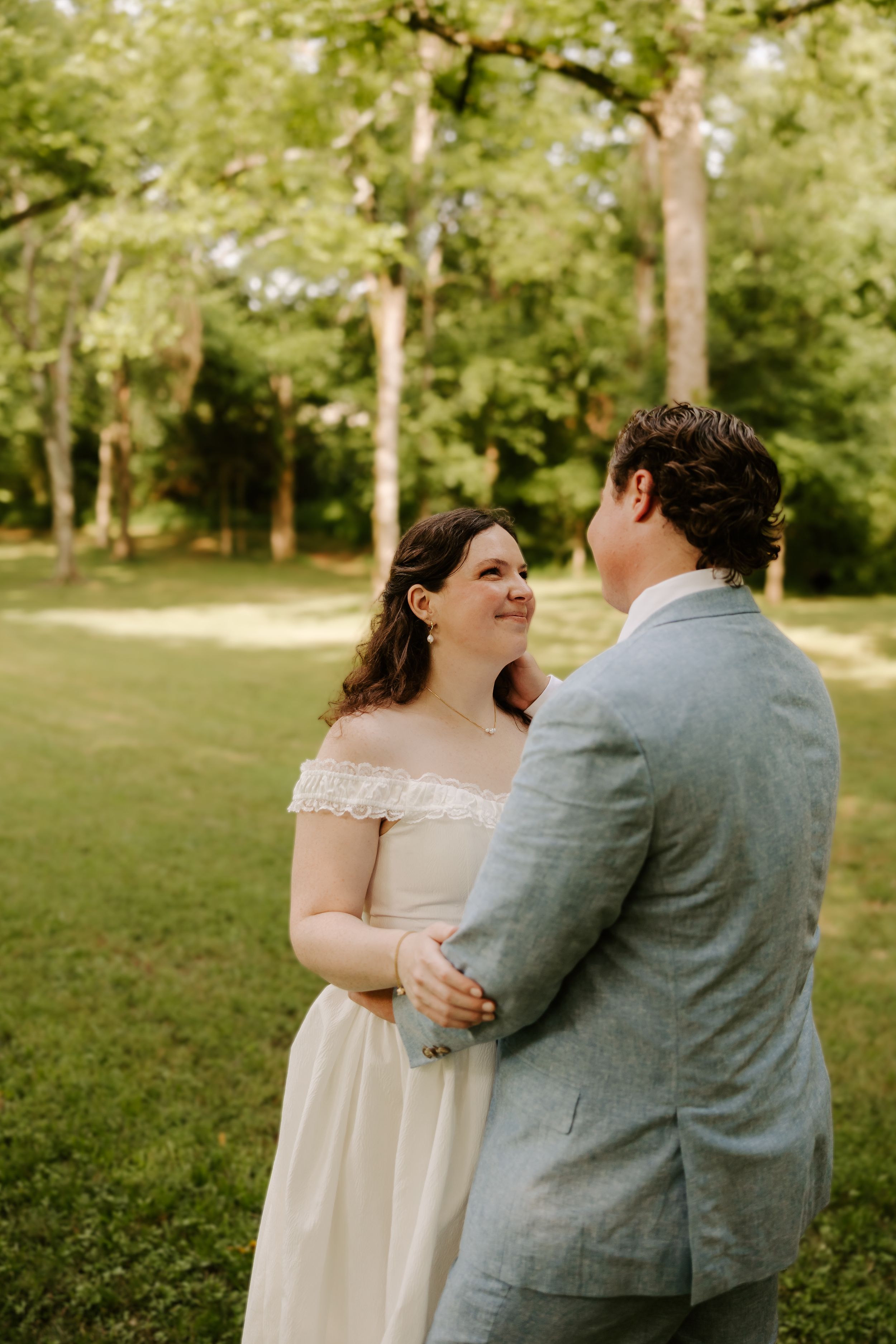 Smiling bride in white off-shoulder gown embracing groom in light blue suit outdoors.
