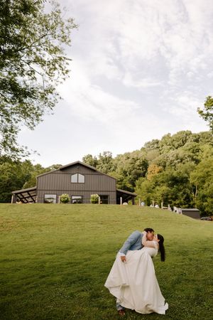 Groom dips bride for a romantic kiss on green lawn with rustic barn wedding venue in background.