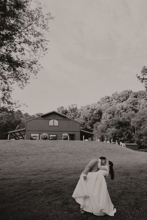 Black and white photo of bride and groom sharing a romantic dip kiss on a green lawn with a barn venue backdrop.