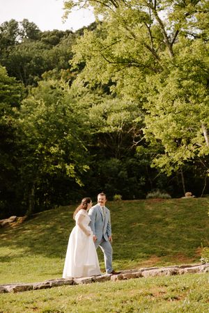 Bride and groom walk hand-in-hand on stone path surrounded by vibrant green hillside trees at outdoor wedding.