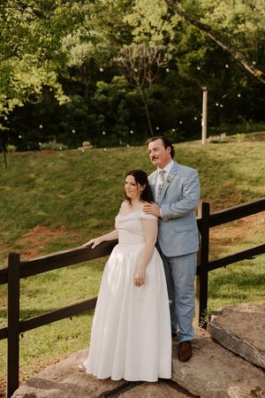 Bride and groom posing by a wooden fence at an outdoor wedding venue surrounded by lush green trees.