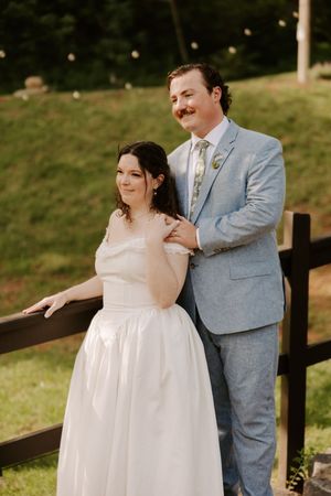Bride and groom pose together by a wooden fence on a green hillside, groom in light blue suit.