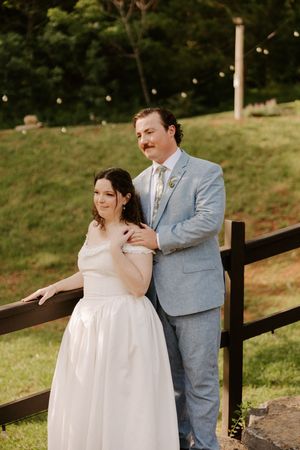 Wedding couple standing together by a rustic fence, groom in light blue suit embracing bride in white gown.