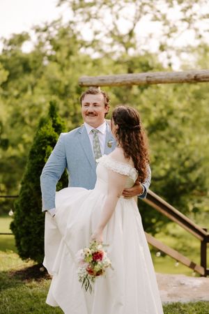 Smiling groom in blue suit lifting bride holding floral bouquet near rustic wooden arbor.
