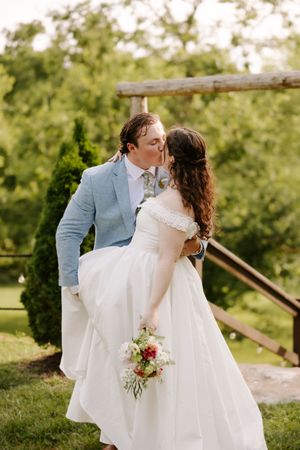 Bride and groom share a romantic kiss outdoors, bride holding colorful bouquet in white off-shoulder gown and groom in light blue suit.
