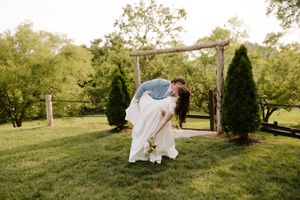 Groom dips bride in white gown near rustic wooden arch flanked by evergreen trees at wedding.