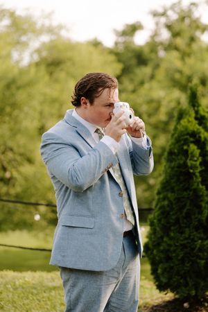Groom in light blue suit photographing with instant camera in outdoor wedding setting.