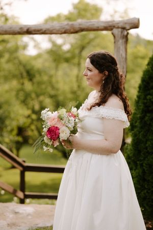 Bride in white off-shoulder gown holding pink and white bouquet under wooden arch.